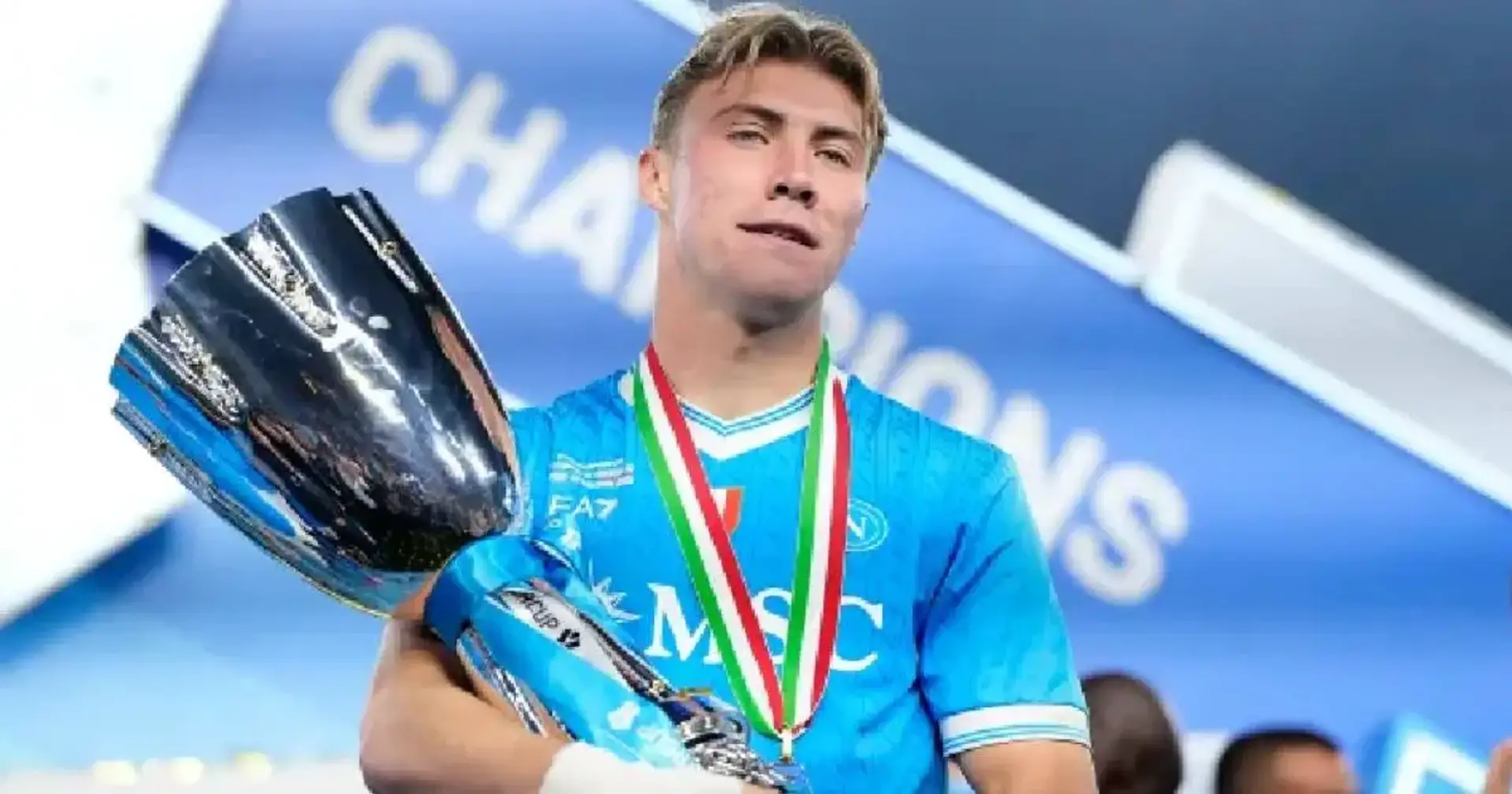 Rasmus Hojlund w błękitnej koszulce sportowej Napoli z medalem w barwach włoskiej flagi na szyi, trzymający w ramionach srebrne trofeum o kielichowatym kształcie. W tle widoczny jest fragment napisu „CHAMPIONS” na niebieskim panelu.